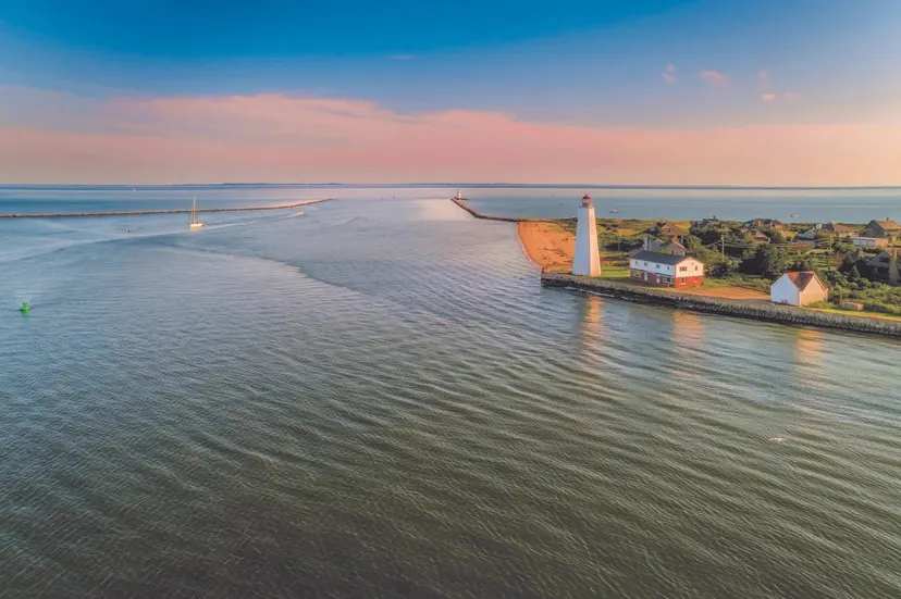 A lighthouse at the mouth of a calm harbor at sunset, with a sailboat heading in